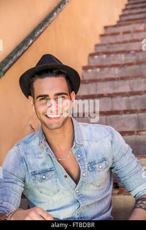 Portrait of handsome man with tattoo sur bras assis sur des escaliers de ciment tout en s'appuyant sur le mur, smiling Banque D'Images