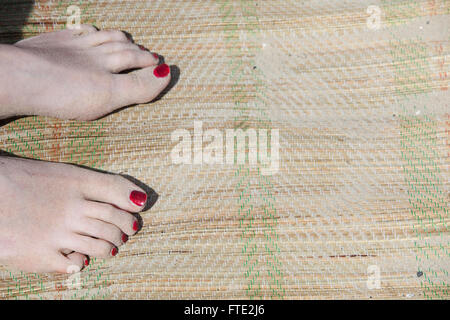 Les ongles des orteils peints en rouge sur un tapis tissé reed avec sable indiquant que la personne est assise sur une plage et est probablement en vacances ou vacances. Banque D'Images