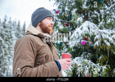 Jeune homme barbu sourire pensif, debout près de l'arbre de Noël décoré et boire du café en plein air Banque D'Images