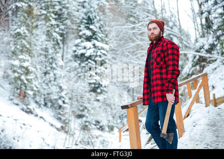 Beau jeune homme pensif avec ax debout dans la forêt d'hiver la montagne et le choix d'un arbre Banque D'Images