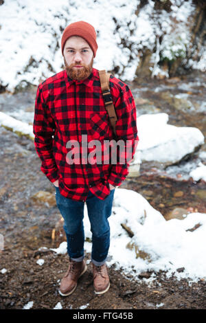 Vue de dessus du beau jeune homme barbu avec sac à dos, debout dans la forêt près de la rivière de montagne d'hiver Banque D'Images