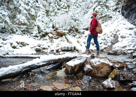Beau jeune homme barbu en chemise à carreaux et jeans avec sac à dos, debout près de la rivière dans la forêt d'hiver Banque D'Images