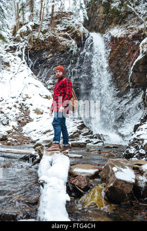 Portrait de jeune homme barbu attrayant avec sac à dos, debout près de cascade en montagne en hiver Banque D'Images