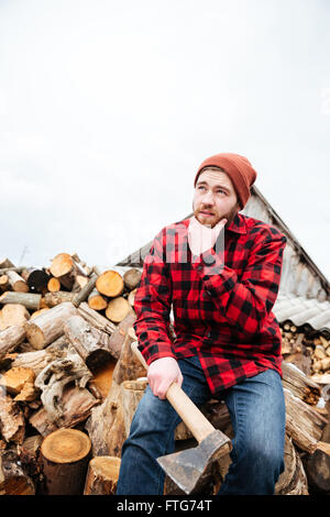 Jeune homme barbu pensif en chemise à carreaux et un chapeau avec ax sitting on logs et la pensée Banque D'Images