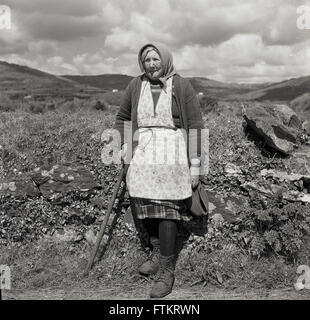 1950s, photo historique par J Allan Cash d'une femme irlandaise mûre portant un tablier ou une robe chasuble sur une jupe en laine, un gilet et une écharpe sur sa tête, debout avec un bâton, par un hedgerow, en Irlande rurale. Banque D'Images