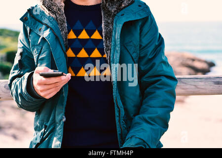 Libre d'un jeune homme portant un manteau de race blanche à l'aide d'un smartphone à l'extérieur, avec l'océan en arrière-plan Banque D'Images