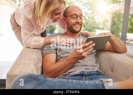 Heureux femme debout derrière son mari assis de canapé dans la salle de séjour, à la fois à la recherche d'une tablette numérique en souriant. Banque D'Images