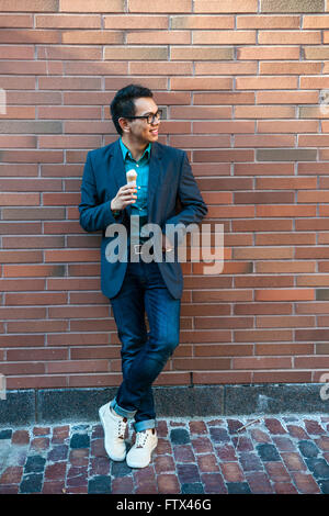 Jeune homme asiatique d'une vingtaine d'années tout en étant debout près de mur de brique à l'extérieur sur une pause à la recherche sur le côté Banque D'Images