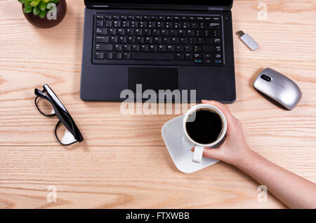 Vue de dessus de hand holding Coffee cup with laptop, bébé plante, souris, clé usb, et lunettes de lecture sur le bureau. Banque D'Images