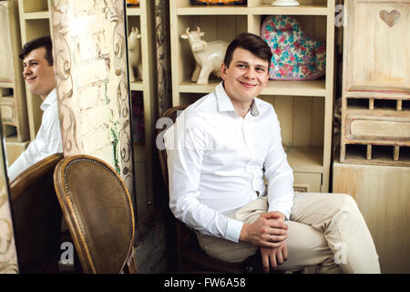 Emotive portrait of smiling handsome crazy hipster en chemise blanche assise sur une chaise vintage avec un smartphone en style rétro Banque D'Images