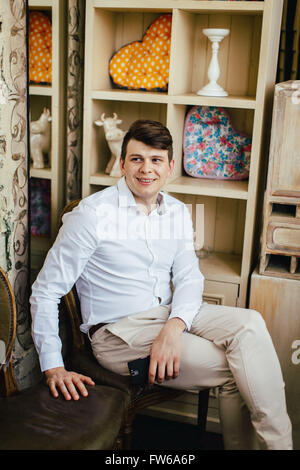 Emotive portrait of smiling handsome hipster en blanc chemise décontractée assis sur une chaise avec un smartphone vintage style rétro en j Banque D'Images