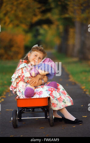 Young Girl lying une poupée de chiffon tout en restant assis dans un chariot rouge Banque D'Images