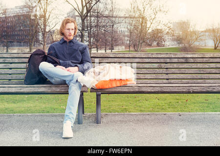 Jeune homme aux longs cheveux rouges et des verres assis dehors sur un banc avec ses sacs Banque D'Images