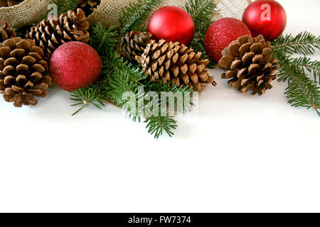 Ornements d'arbre de Noël rouge, pommes de pin, branches de conifères, et tissu jute créent un cadre, isolé sur une case white Banque D'Images