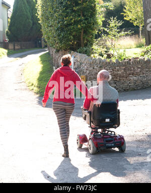 Vue arrière d'un homme âgé à l'aide d'un scooter de mobilité avec son soignant marcher à côté de lui Banque D'Images