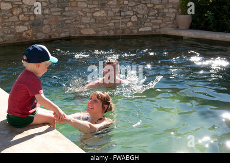 Tout-petit assise sur le bord d'une piscine avec ses parents l'encourageant à aller dans Banque D'Images