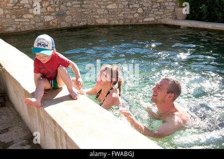 Tout-petit assise sur le bord d'une piscine avec ses parents debout dans la piscine Banque D'Images