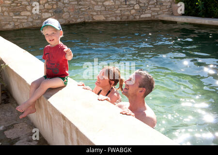 Tout-petit assise sur le bord d'une piscine avec ses parents debout dans la piscine Banque D'Images