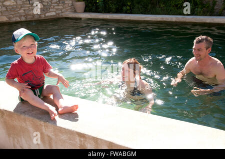 Tout-petit assise sur le bord d'une piscine avec ses parents debout dans la piscine Banque D'Images