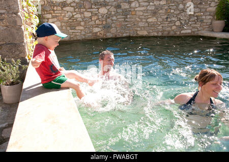 Tout-petit assise sur le bord d'une piscine éclaboussant ses parents qui sont debout dans la piscine Banque D'Images