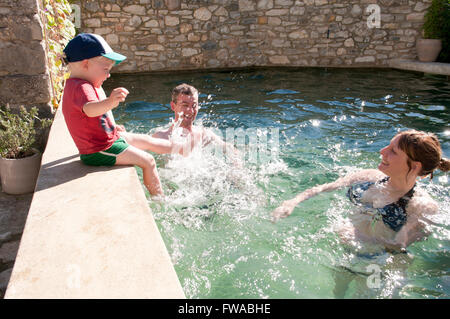 Tout-petit assise sur le bord d'une piscine éclaboussant ses parents qui sont debout dans la piscine Banque D'Images