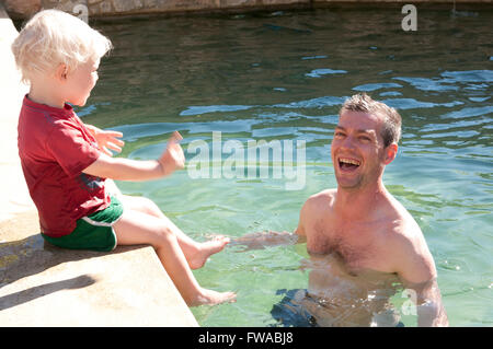 Tout-petit assise sur le bord d'une piscine avec son père debout dans la piscine de rire Banque D'Images