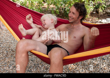 Père et fils rire Être souriant, espiègle, assis dans un hamac rouge et jaune Banque D'Images