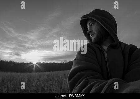 Self portrait noir et blanc d'un homme debout, dans un champ à l'extérieur regardant par-dessus son épaule vers le soleil. Banque D'Images