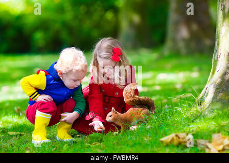 Kids feeding écureuil dans le parc de l'automne. Petit garçon et fille en robe rouge et des bottes de pluie regarder les animaux sauvages dans la forêt d'automne Banque D'Images