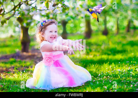 Enfant jouant avec un oiseau. Heureux rire petite fille en costume de fée avec des ailes de nourrir un perroquet dans un cerisier jardin Banque D'Images