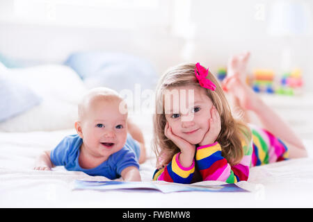 Funny happy girl reading a book et jouer avec bébé nouveau-né garçon au lit. Les enfants jouent à la maison. Pépinière blanc. Banque D'Images