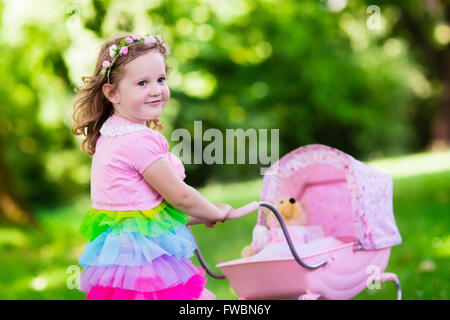 Petite fille poussant poussette jouet avec l'ours. Tout-petit enfant en robe rose jouant à la poupée buggy. Kids Birthday party. Banque D'Images