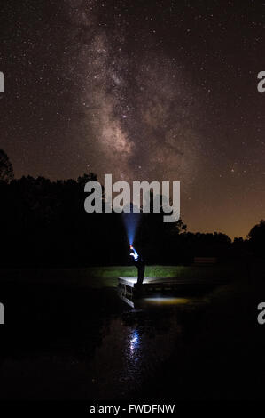 Un homme debout sur le pont au-dessus d'un plan d'eau la nuit qui brille une lampe vers la voie lactée. Banque D'Images