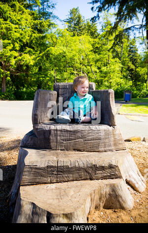 Un jeune garçon est assis sur une souche sculptée dans un siège à un parc à l'extérieur pour ce mignon et simple portrait. Banque D'Images