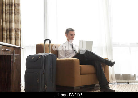 Portrait of handsome young man wearing white shirt officiel cravate noire et assis dans un fauteuil dans chambre d'hôtel avec paniers Banque D'Images