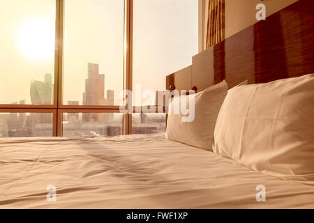 Femme de chambre lit avec oreillers blanc propre et les draps de lit dans la chambre vide. Close-up. Flair de l'objectif à la lumière du soleil. Lever du Soleil vue sur la ville Banque D'Images