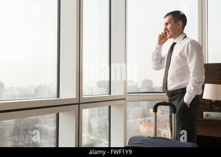 Young happy traveler businessman wearing chemise blanche et cravate faisant appel après son arrivée dans la chambre d'hôtel avec son assurance Banque D'Images