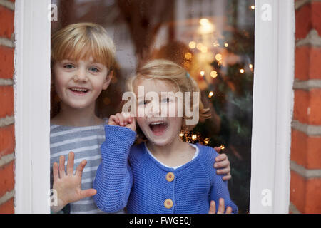 Deux enfants, un garçon et une fille, regarder par une fenêtre à la maison avec des expressions excité. Banque D'Images