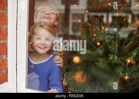 Deux enfants, un garçon et une fille, regarder par une fenêtre à la maison à côté d'un arbre de Noël. Banque D'Images