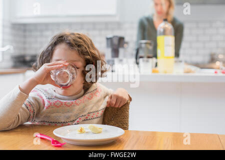 Un enfant assis à une table dans un verre d'alcool, une femme debout derrière elle dans une cuisine domestique. Banque D'Images