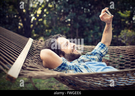 Un homme étendu dans un hamac de jardin en tenant vos autoportraits avec son téléphone. Banque D'Images