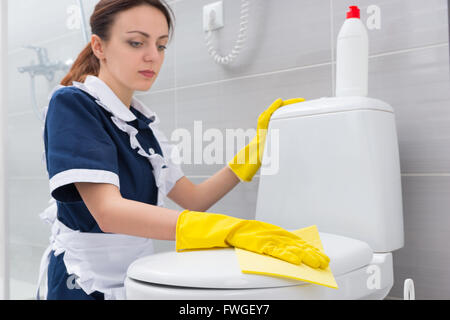 Femme de ménage ou dans une salle de bains blanche un nettoyage uniforme à genoux l'essuyage toilettes avec un chiffon jaune avec des gants. Banque D'Images