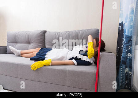 Femme de ménage fatigué ou en uniforme de détente sur le travail étendu sur un canapé couvrant ses yeux pour une sieste avec son mop aux côtés Banque D'Images