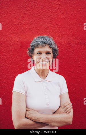 Portrait of attractive young woman with her arms crossed standing contre fond rouge. Smiling mature femme contre la ré Banque D'Images