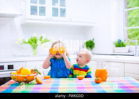 Drôle mignon et adorable petite fille garçon bébé boire du jus d'orange fraîchement pressé pour petit-déjeuner sain dans une cuisine blanche Banque D'Images