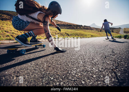 Jeune femme patinage avec son ami en arrière-plan. Les jeunes longboard on rural road sur journée d'été. Banque D'Images