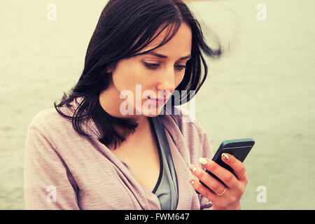 Une jeune femme textos sur téléphone cellulaire contre l'eau , focus sélectif, image colorisée Banque D'Images