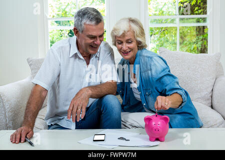 Senior woman putting coins in piggy bank lors d'un entretien avec l'homme sur canapé Banque D'Images