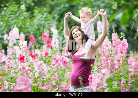 Les jeunes et mettre en place mère enceinte active jouant avec sa petite fille dans un jardin avec des fleurs roses Banque D'Images