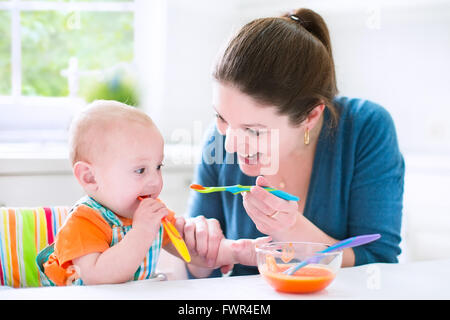 Young attractive mère allaiter son bébé mignon fils, lui donnant son premier aliment solide, saine de pure végétale carotte Banque D'Images